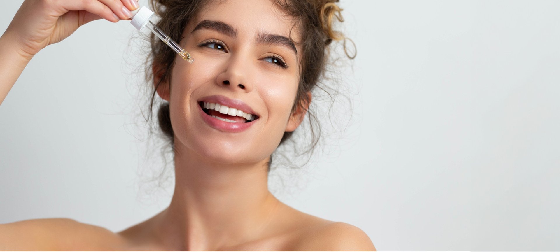 Eine lächelnde junge Frau tröpfelt sich ein Serum mit einer Pipette auf ihre rechte Wange