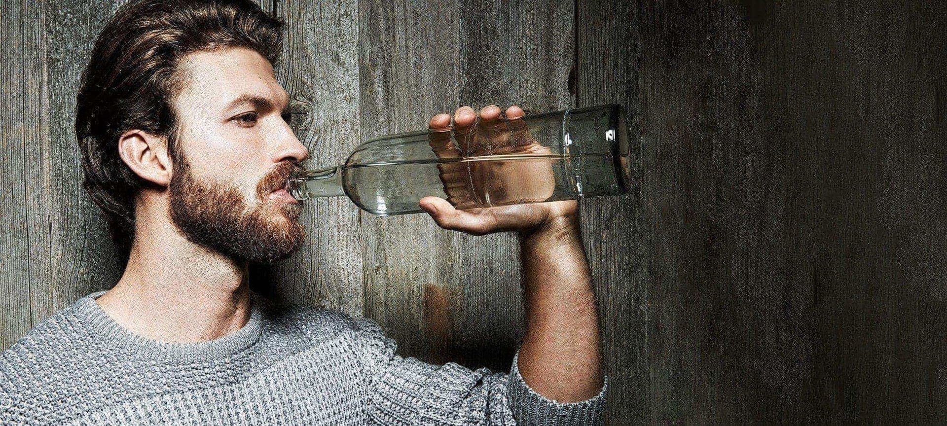 Bärtiger Mann trinkt aus Wasserfalsche vor einer Holzwand