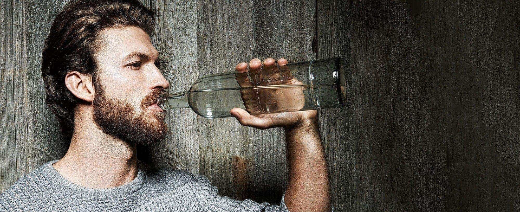 Bärtiger Mann trinkt aus Wasserfalsche vor einer Holzwand