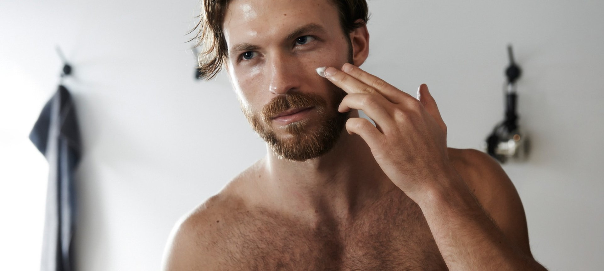 Ein junger Mann betrachtet sich im Spiegel und trägt dabei auf seine Wange eine Creme auf