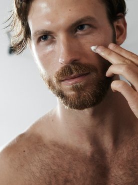 Ein junger Mann betrachtet sich im Spiegel und trägt dabei auf seine Wange eine Creme auf