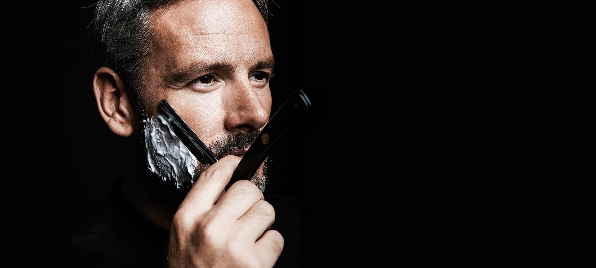 Mann vor schwarzem Hintergrund rasiert sich mit einem Rasiermesser