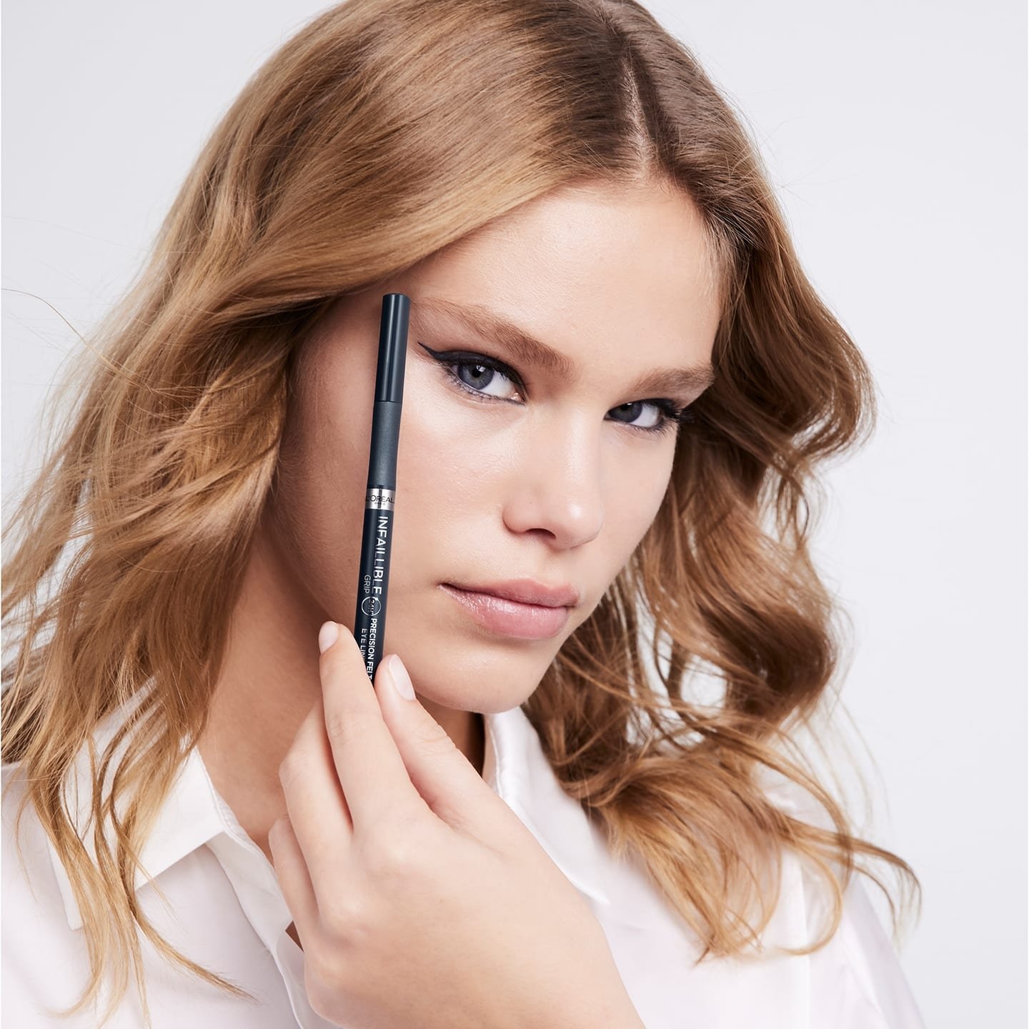 Blonde Frau hält Infaillible Eyeliner Infaillible 27h Grip Precision Felt Liner 01 schwarz Produkt neben ihr Gesicht